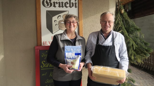Ein Mann und eine Frau stehen vor dem Käseladen. Er hat einen Laib in der Hand, sie präsentiert ein Fondue. Ein Mann und eine Frau stehen vor dem Käseladen. Er hat einen Laib in der Hand, sie präsentiert ein Fondue.