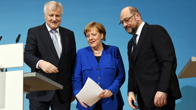 Drei Personen in einer Reihe vor einem Rednerpult stehend. Drei Personen in einer Reihe vor einem Rednerpult stehend.