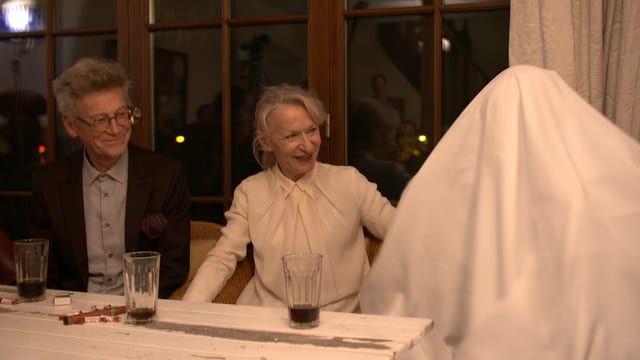 Ein Mann und eine Frau sitzen an einem Tisch, daneben ein Mann mit einem Tischtuch auf dem Kopf.