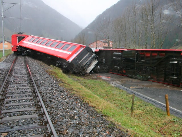 Links Eisenbahnschiene, rechts liegt ein roter Eisenbahnwagen quer zur Schiene, ein zweiter sogar auf der benachbarten Strasse einige Meter vom Gleis entfernt. Links Eisenbahnschiene, rechts liegt ein roter Eisenbahnwagen quer zur Schiene, ein zweiter sogar auf der benachbarten Strasse einige Meter vom Gleis entfernt.