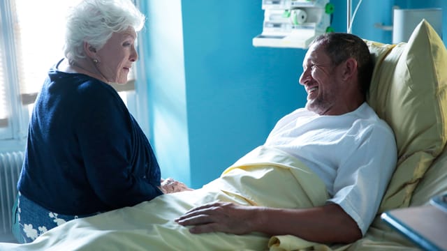 Alte Frau sitzt im Spital am Bettrand, im Bett ein Mann. Alte Frau sitzt im Spital am Bettrand, im Bett ein Mann.