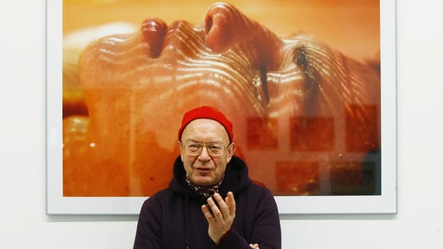älterer Herr mit roter Mütze steht vor einer grossen Fotografie in Orangetönen. älterer Herr mit roter Mütze steht vor einer grossen Fotografie in Orangetönen.