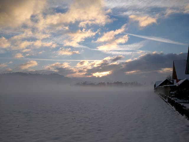 Über einem verschneiten Feld liegt eine seichte Bodennebelschicht. Der Himmel ist teilweise bewölkt, teilweise klar. Am Horizonte macht sich hinter den Wolken die Sonne bemerkbar.
