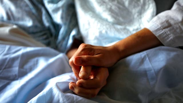 Die Hand eines Arztes hält eine Patientenhand auf den weissen Laken eines Spitalbetts. Die Hand eines Arztes hält eine Patientenhand auf den weissen Laken eines Spitalbetts.