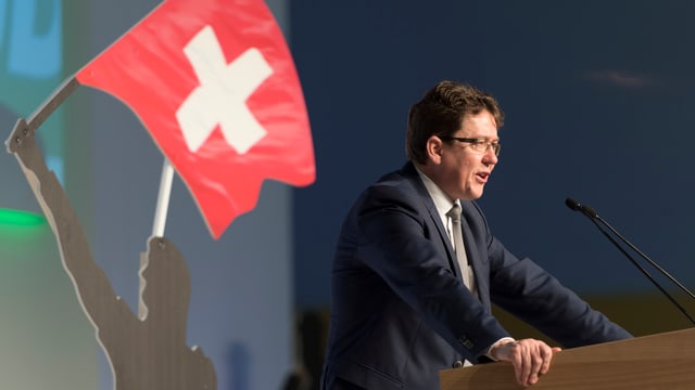 Albert Rösti an einem Rednerpult von der Seite. Am Rand ist eine Figur mit einer Schweizer-Fahne. Albert Rösti an einem Rednerpult von der Seite. Am Rand ist eine Figur mit einer Schweizer-Fahne.