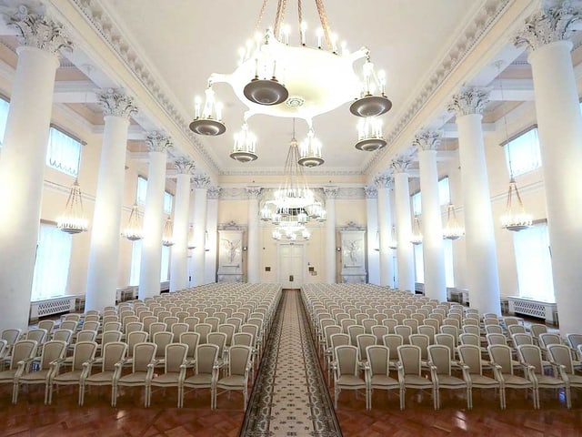 Der weisse Ballsaal vom Smolny-Institut: Weisse Stühle sind symmetrisch angeordnet, in der Mitte liegt ein langer Teppich, der bis zur Türe reicht. Der weisse Ballsaal vom Smolny-Institut: Weisse Stühle sind symmetrisch angeordnet, in der Mitte liegt ein langer Teppich, der bis zur Türe reicht.