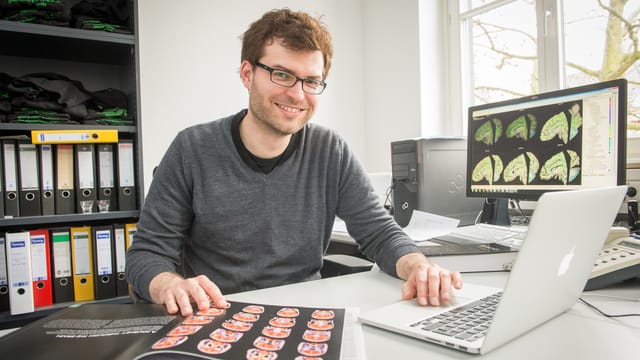 Sebastian Markett hinter seinem Schreibtisch mit Buch und Laptop. Sebastian Markett hinter seinem Schreibtisch mit Buch und Laptop.