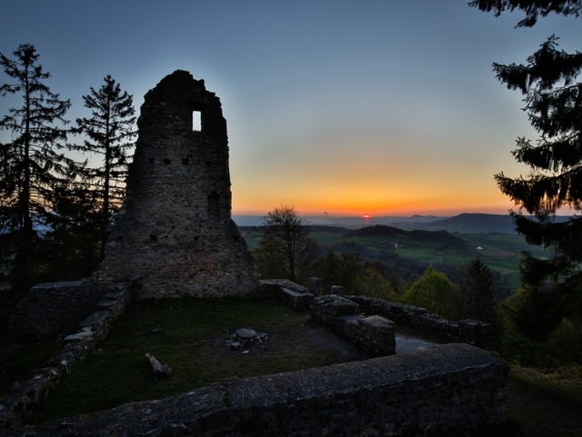 Im Vordergrund eine Burgruine. Im Hintergrund geht am Horizont die Sonne auf und verfärbt den Himmel rötlich.