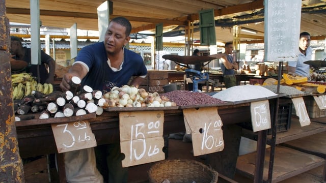 Ein dunkler kubanischer Verkäufer steht hinter einem Markstand. Vor ihm liegen diverse Produkte (zum Beispiel Manjok, Zwiebeln und Bananen) Ein dunkler kubanischer Verkäufer steht hinter einem Markstand. Vor ihm liegen diverse Produkte (zum Beispiel Manjok, Zwiebeln und Bananen)