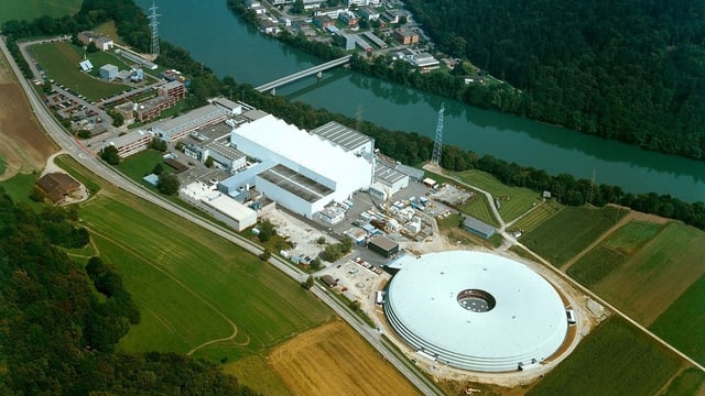 Aufnahme aus der Luft vom Paul Scherrer Institut. Zu sehen ist ein grosses rundes, flaches Gebäude. Daneben ein hohes, industrieartiges weisses Gebäude. Rund um die Anlage hat es Ackerland. Aufnahme aus der Luft vom Paul Scherrer Institut. Zu sehen ist ein grosses rundes, flaches Gebäude. Daneben ein hohes, industrieartiges weisses Gebäude. Rund um die Anlage hat es Ackerland.