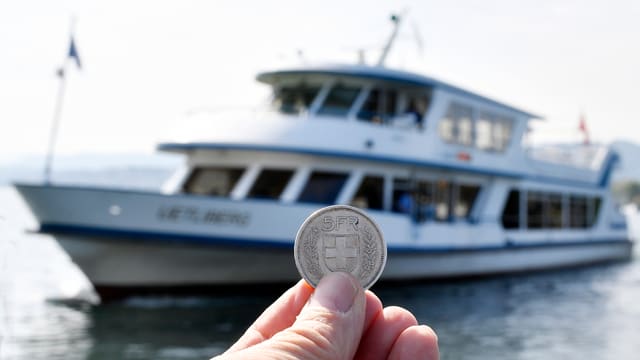 Eine Hand hält eine Fünf-Franken-Münze vor einem Schiff in die Kamera. Eine Hand hält eine Fünf-Franken-Münze vor einem Schiff in die Kamera.