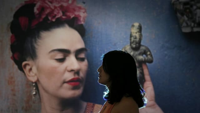 Frau schaut über den linken Bildrand heraus, im Hintergrund ein grosses Abbild von Frida Kahlo. Frau schaut über den linken Bildrand heraus, im Hintergrund ein grosses Abbild von Frida Kahlo.