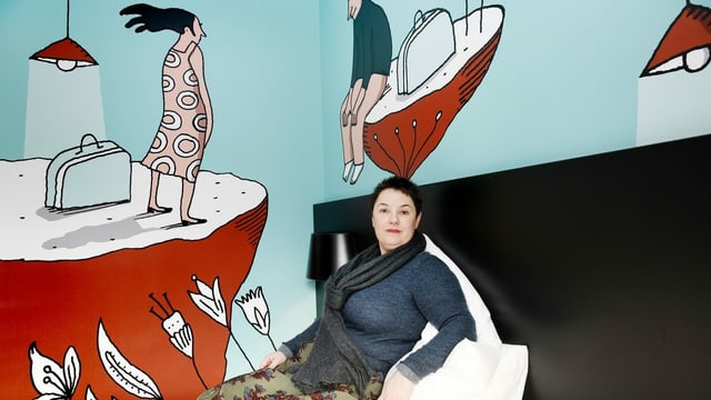 eine Frau sitzt auf einem Bett, dahinter eine bemalte Wand eine Frau sitzt auf einem Bett, dahinter eine bemalte Wand