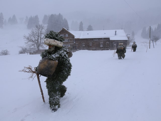 Ein Silvesterklaus läuft durch den Schnee. Im Hintergrund weitere Kläuse und ein Bauernhaus.