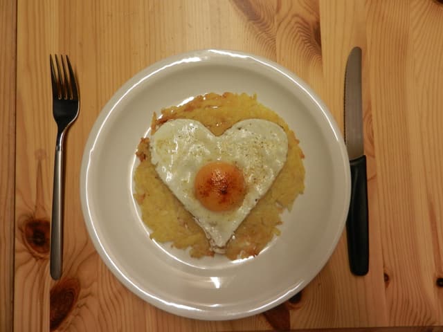 Ein Teller auf einem Holztisch, darauf Rösti und ein Spiegelei in Herzform.