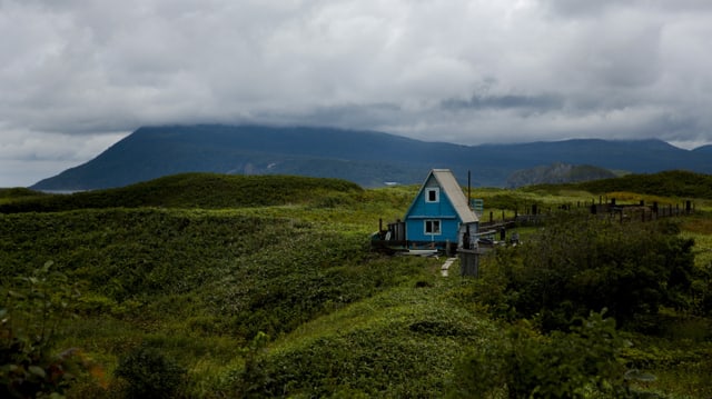 Grüne, hügelige Landschaft, darin steht ein blaues Holzhäuschen. Grüne, hügelige Landschaft, darin steht ein blaues Holzhäuschen.