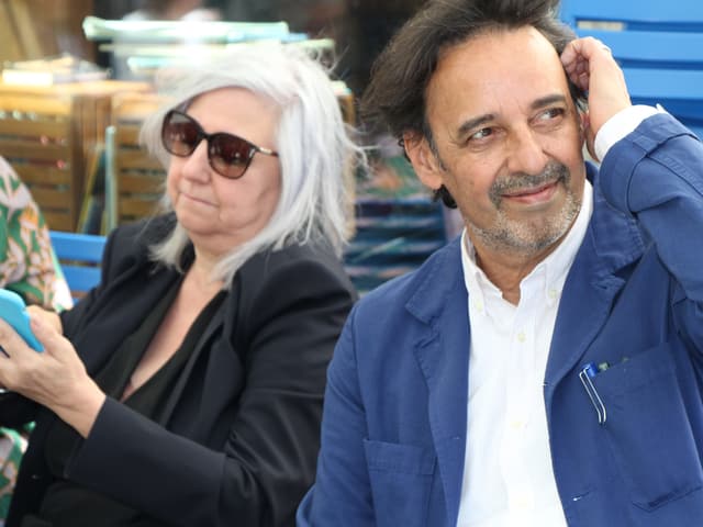 Ein Mann mit weissem Hemd, blauem Jackett und braunen Haaren. Links von ihm eine Frau mit langen grauen Haaren, Sonnenbrille, schaut aufs Handy. Ein Mann mit weissem Hemd, blauem Jackett und braunen Haaren. Links von ihm eine Frau mit langen grauen Haaren, Sonnenbrille, schaut aufs Handy.