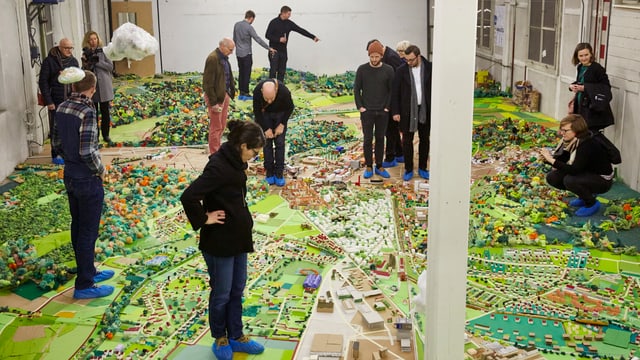 buntes Stadtmodell in einem grossen Raum, darauf stehen Leute buntes Stadtmodell in einem grossen Raum, darauf stehen Leute