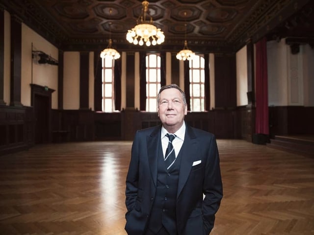 Mann steht in grossem Saal mit Parkett, Kronleuchtern und roten Vorhängen. Mann steht in grossem Saal mit Parkett, Kronleuchtern und roten Vorhängen.