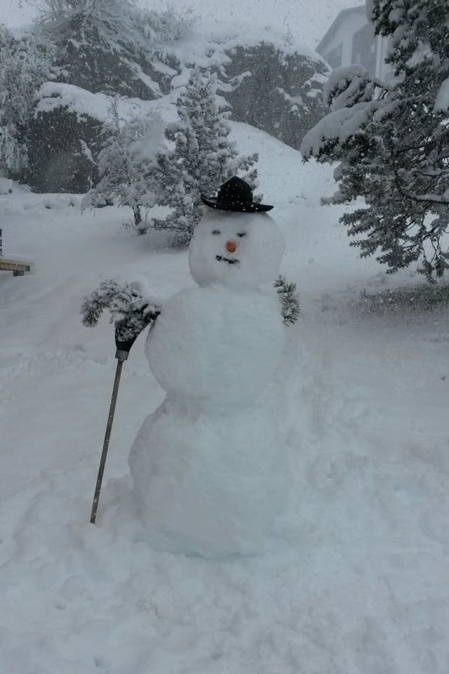 Auf den Bäumen, der Wiese und den Häusern rundum liegen 15 bis 20 Zentimeter Schnee. In der Mitte steht ein grosser Schneemann mit einem Hut, einer Rübe und einem Besen.