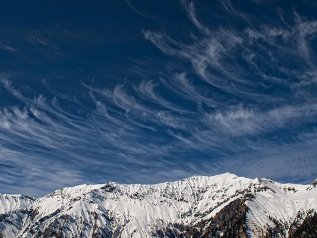 Blauer Himmel mit feinen, haarartigen Wolken. Sie haben eine hackenform. Unten Schneeberge.