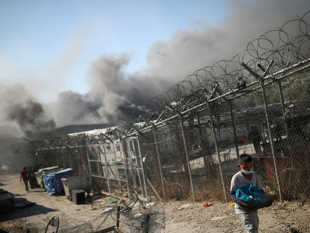 Ein Junge geht mit seinem Hab und Gut in den H&auml;nden den Zaun des Lagers entlang, dahinter sind Rauchschwaden zu sehen.