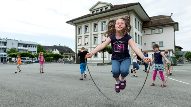 Ein Mädchen beim Seilspringen auf einem Pausenplatz. Im Hintergrund ein Schulhaus. Ein Mädchen beim Seilspringen auf einem Pausenplatz. Im Hintergrund ein Schulhaus.