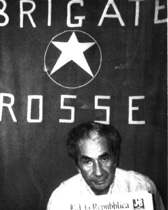 EIn älterer Mann blickt in die Kamera, dahinter steht "Brigate Rosse" an der Wand. EIn älterer Mann blickt in die Kamera, dahinter steht "Brigate Rosse" an der Wand.