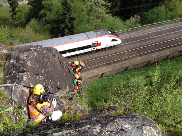 Kameramann hängt oben im Felsen, unten sind zwei weitere Personen angeseilt. Direkt darunter eine Bahnschiene mit einem vorbeifahrenden Zug.
