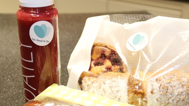 Ein frisch gepresster Saft und Backwaren mit einem Kleber von "Too Good To Go" darauf. Ein frisch gepresster Saft und Backwaren mit einem Kleber von "Too Good To Go" darauf.