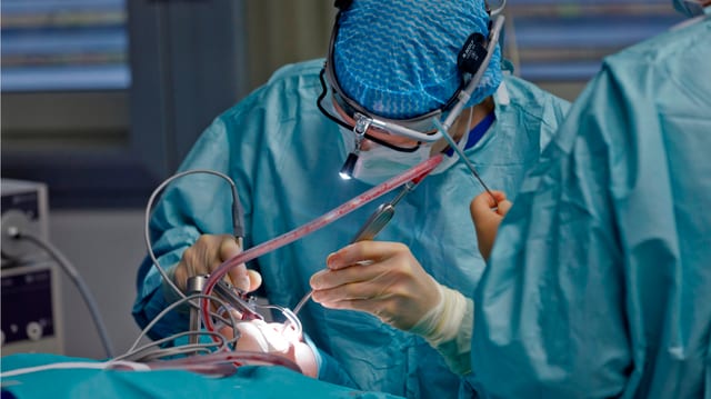 Ein Arzt in blau-grünem Überkleid operiert in einem OP eine Person am Kopf.
