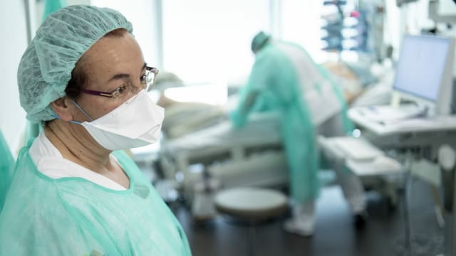 Pflegerin im Vordergrund, im Hintergrund eine Pflegeperson arbeitet mit einem Patienten, beide tragen Gesichtsmasken. Pflegerin im Vordergrund, im Hintergrund eine Pflegeperson arbeitet mit einem Patienten, beide tragen Gesichtsmasken.