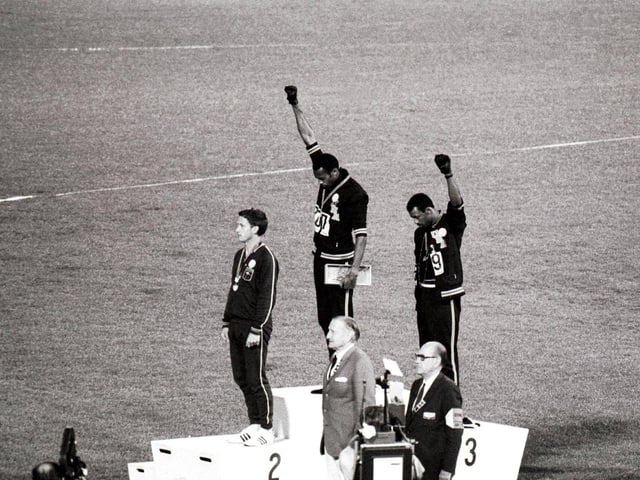 Tommie Smith (Mitte) und John Carlos (rechts) mit hochgereckter Faust. Tommie Smith (Mitte) und John Carlos (rechts) mit hochgereckter Faust.