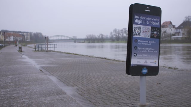 Ein Smartphone so gross wie ein Mensch am Flussufer von Monden. Es wirbt für die «Beacon-Mile». Ein Smartphone so gross wie ein Mensch am Flussufer von Monden. Es wirbt für die «Beacon-Mile».