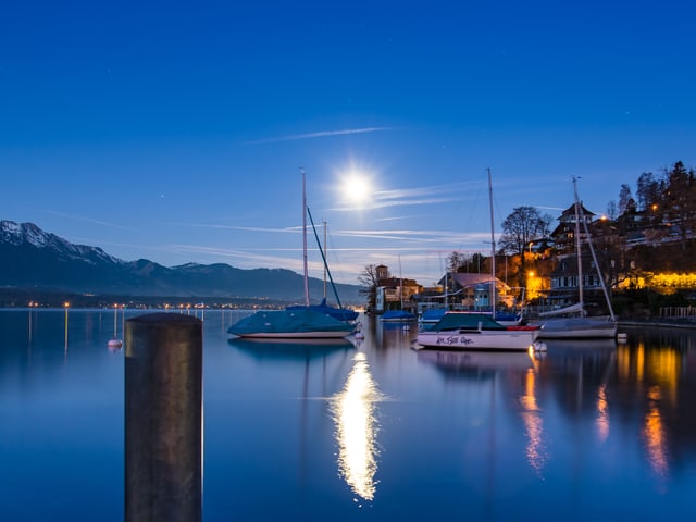 Der Himmel ist tiefblau und praktisch wolkenlos, tief am Himmel scheint der Mond. Im Vordergrund der Thunersee, ziemlich glatt. Am rechten Bildrand einige Häuser und Lichter.