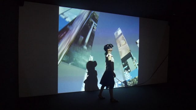 Eine Frau mit VR-Brille steht in einem dunklen Raum. Auf ihren Körper und an die Wand wird ein Bild projiziert, auf dem Wolkenkratzer zu sehen sind. Eine Frau mit VR-Brille steht in einem dunklen Raum. Auf ihren Körper und an die Wand wird ein Bild projiziert, auf dem Wolkenkratzer zu sehen sind.
