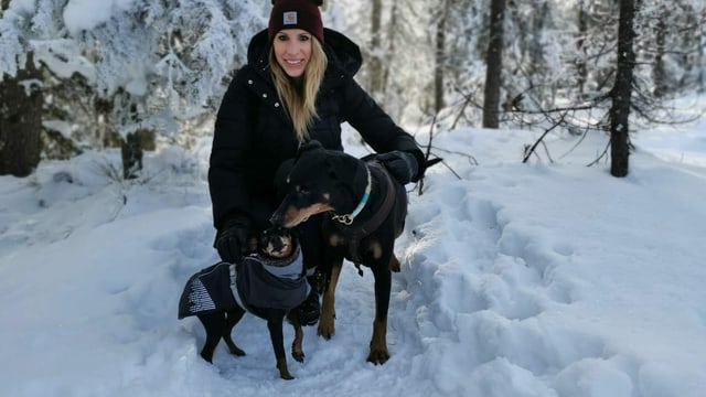 Eine junge Frau in schwarzen Winterkleidern ist mit zwei Hunden im verschneiten Wald unterwegs. Eine junge Frau in schwarzen Winterkleidern ist mit zwei Hunden im verschneiten Wald unterwegs.