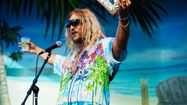 Mann auf Bühne im Hawaihemd mit Buch und Drink in der Hand. Mann auf Bühne im Hawaihemd mit Buch und Drink in der Hand.