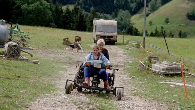 Laura und Thom fahren auf einem Rasenmäher einen Feldweg hinauf.