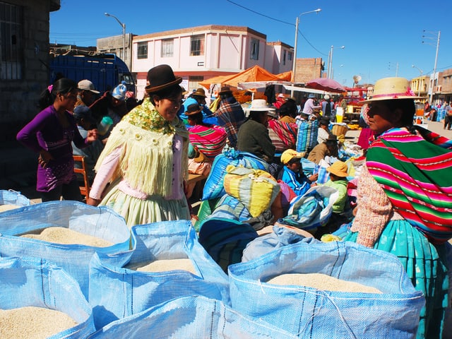 im vordergrund sind grosse blaue Säcke voll mit hellen Körnchen zu sehen, im Hintergrund ein bunter Markt.