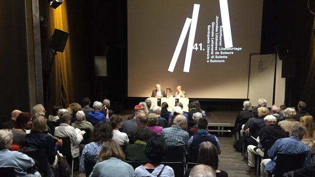 Ein Saal, gefüllt mit Menschen, die dem Schriftsteller Ernst Halter, ganz klein am Ende des Saales, zuhören. Ein Saal, gefüllt mit Menschen, die dem Schriftsteller Ernst Halter, ganz klein am Ende des Saales, zuhören.