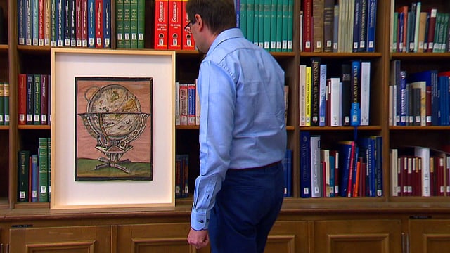 Mann vor einem eingerahmten Pergament stehend, Dahinter steht ein Regal mit Büchern. Mann vor einem eingerahmten Pergament stehend, Dahinter steht ein Regal mit Büchern.