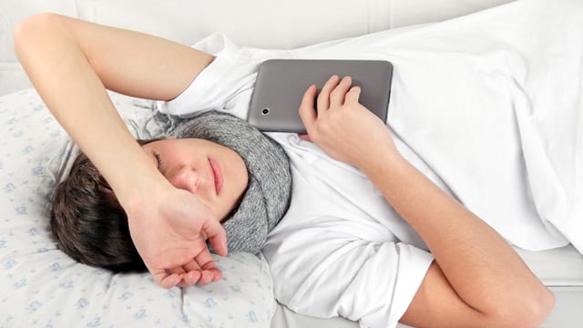 Ein Mann liegt mit geschlossenen Augen im Bett, einen grauen Schal um den Hals gebunden und ein graues Tablet in der Hand. Ein Mann liegt mit geschlossenen Augen im Bett, einen grauen Schal um den Hals gebunden und ein graues Tablet in der Hand.