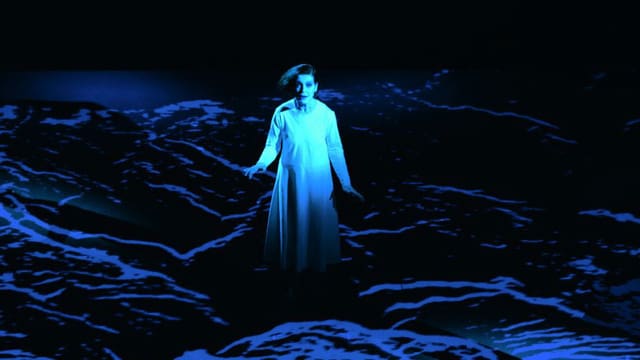 Bühnenbild: Ein in weiss gekleidetes Mädchen steht in einer dunklen Landschaft.
