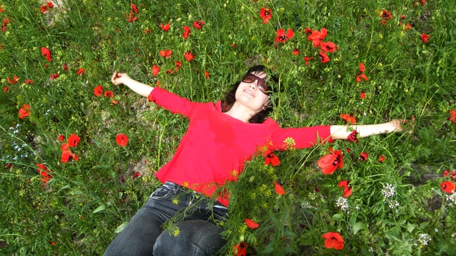 Eine Frau liegt strahlend und glücklich auf einer Wiese, aus der rote Blüten wachsen. Eine Frau liegt strahlend und glücklich auf einer Wiese, aus der rote Blüten wachsen.
