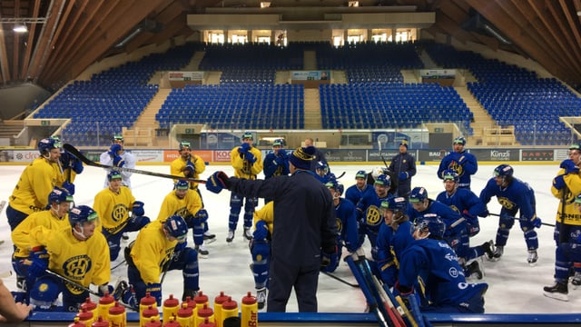 Die Spieler stehen im Halbkreis auf dem Eis um den Trainer und hören ihm zu.