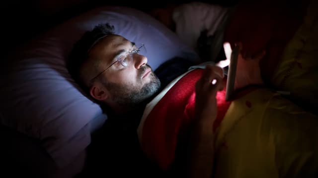 Ein Mann liegt im Bett. Er wird nur durch den Schein seines Smartphones beleuchtet. 