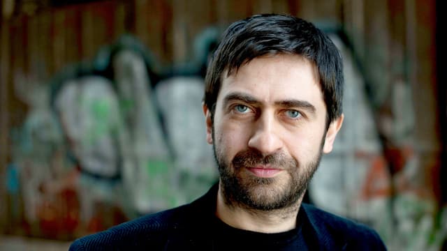 Emin Alper, Porträt nah, vor einer Wand mit Graffiti. Emin Alper, Porträt nah, vor einer Wand mit Graffiti.