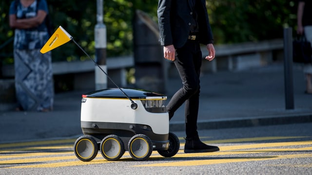 Ein Roboter mit sechs Rädern fährt neben einem Fussgänger über einen Fussgängerstreifen. Ein Roboter mit sechs Rädern fährt neben einem Fussgänger über einen Fussgängerstreifen.