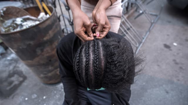 Eine Person flechtet die Haare einer am Boden kauernden Frau. Eine Person flechtet die Haare einer am Boden kauernden Frau.
